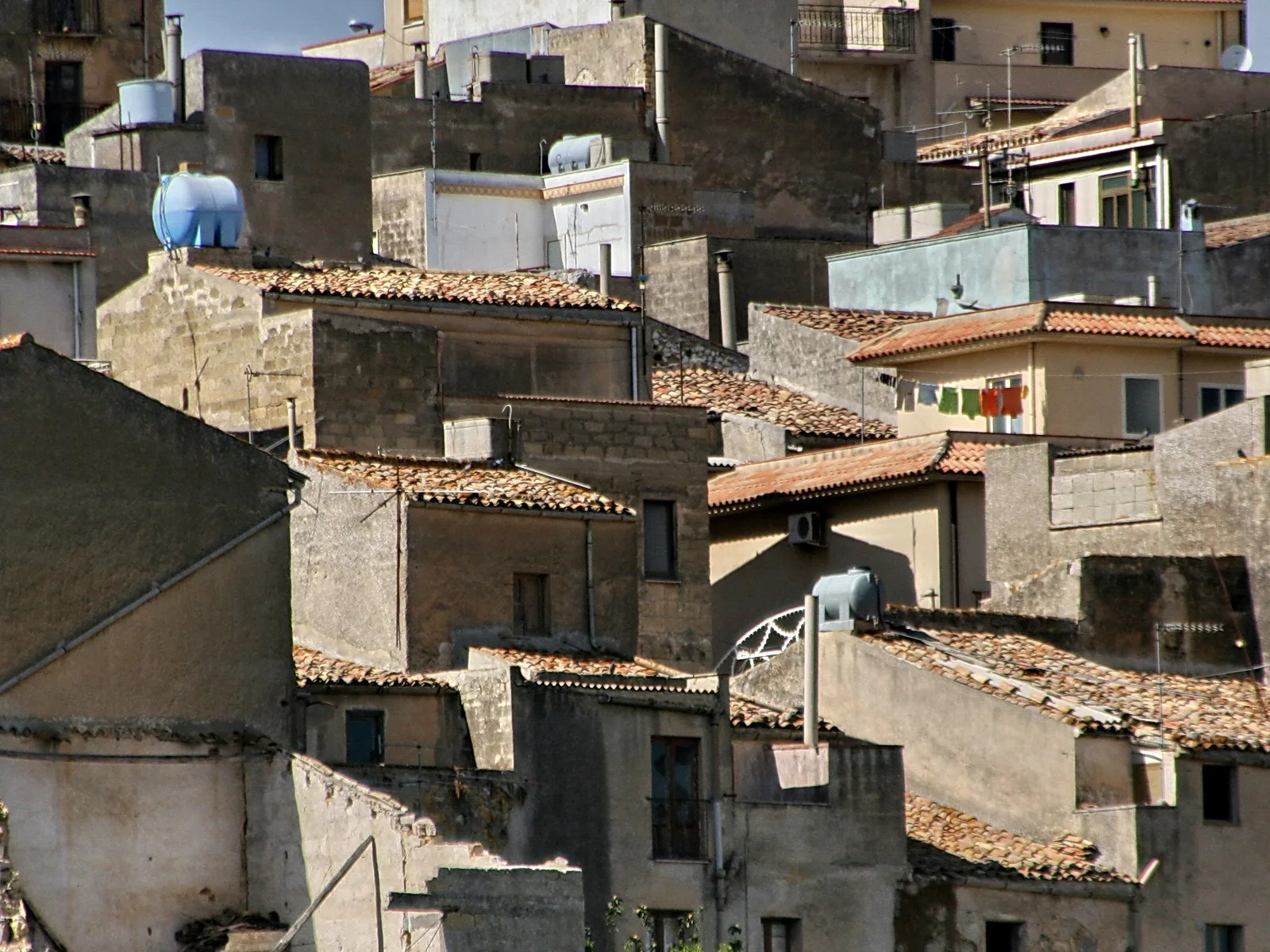 Dicht gedrängte alte Wohnhäuser mit Terrakotta-Dächern, dazwischen ein blauer Wassertank und Wäsche auf einer Leine.