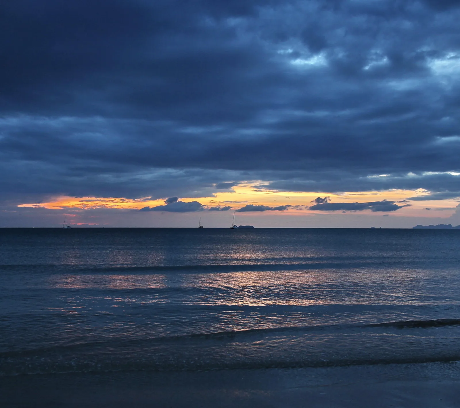 Meer in der Dämmerung unter dunklen Wolken, am Horizont ein orangefarbener Streifen und mehrere Segelboote