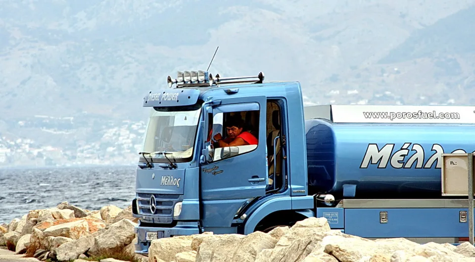 Blauer Tankwagen mit der Aufschrift Mélλος fährt über Felsbrocken an einer Küste, dahinter Meer und hügelige Berge im Dunst. Ein Fahrer sitzt in der Kabine.