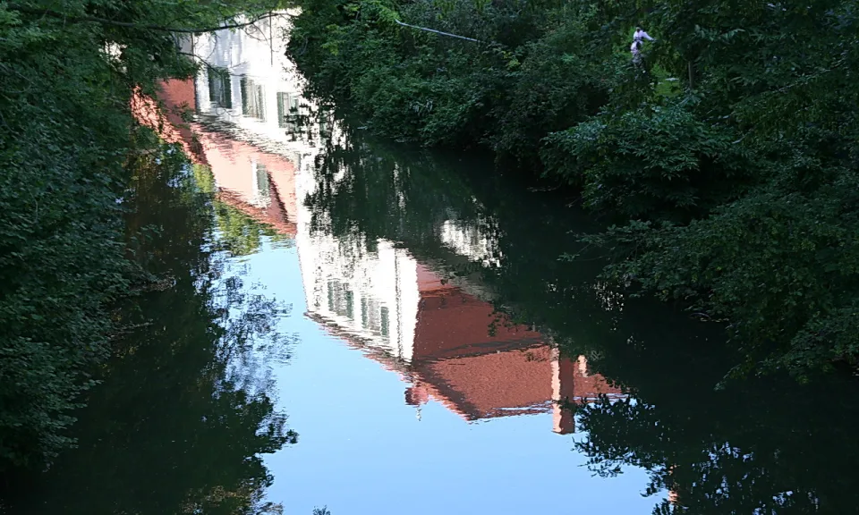Ein Fluss mit dunklem Wasser, in dem sich eine weiße und eine rotbraune Hausfassade spiegeln, eingerahmt von dichtem grünem Gebüsch.