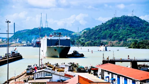Ein großes Frachtschiff mit dunklem Rumpf fährt auf dem Panamakanal, daneben ein Schlepper, im Hintergrund bewaldete Hügel und Strommasten, im Vordergrund Dächer und Kaianlagen.