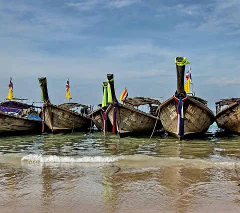 Mehrere hölzerne Longtailboote mit bunten Bändern und thailändischen Flaggen liegen am Strand, davor brechen flache Wellen im seichten Wasser unter einem blauen Himmel mit Schleierwolken.
