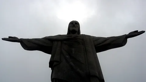 Dunkle Jesusstatue mit weit ausgebreiteten Armen vor bewölktem Himmel, von unten fotografiert