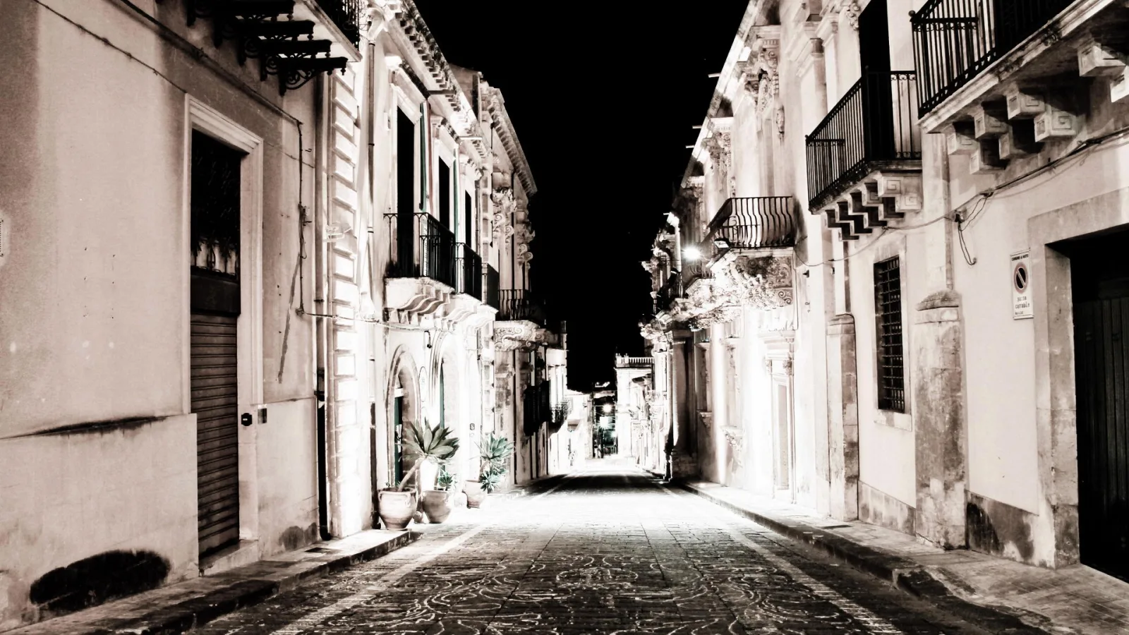 Schmale, leer gepflasterte Gasse bei Nacht, gesäumt von barocken Palazzi mit Balkonen und verzierten Fassaden, Topfpflanzen am linken Bildrand, Laternenlicht auf dem Pflaster.