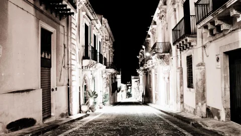 Schmale, leer gepflasterte Gasse bei Nacht, gesäumt von barocken Palazzi mit Balkonen und verzierten Fassaden, Topfpflanzen am linken Bildrand, Laternenlicht auf dem Pflaster.