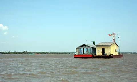 Ein kleines gelb-graues Hausboot mit rotem Rumpf treibt auf braunem Flusswasser, rechts ein Mast mit roter Flagge, links flaches Ufer mit Bäumen unter blauem Himmel.