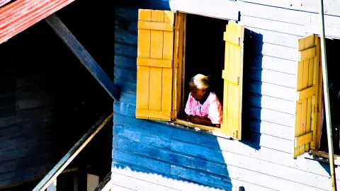 Eine ältere Frau mit weißen Haaren und rosa Bluse lehnt am Fenster eines Holzhauses mit gelben Fensterläden und blau-weißen Holzwänden.