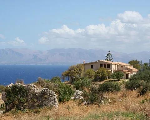 Ein cremefarbenes zweigeschossiges Landhaus mit Ziegeldach steht auf einem trockenen Hügel über dem blauen Meer. Davor liegen helle Felsbrocken und Olivenbäume, im Hintergrund erheben sich bergige Küstenhänge unter einem leicht bewölkten Himmel.