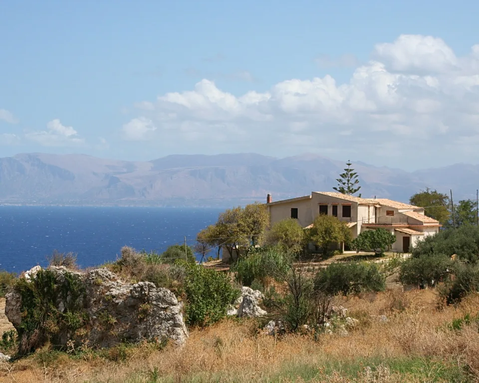 Ein cremefarbenes zweigeschossiges Landhaus mit Ziegeldach steht auf einem trockenen Hügel über dem blauen Meer. Davor liegen helle Felsbrocken und Olivenbäume, im Hintergrund erheben sich bergige Küstenhänge unter einem leicht bewölkten Himmel.