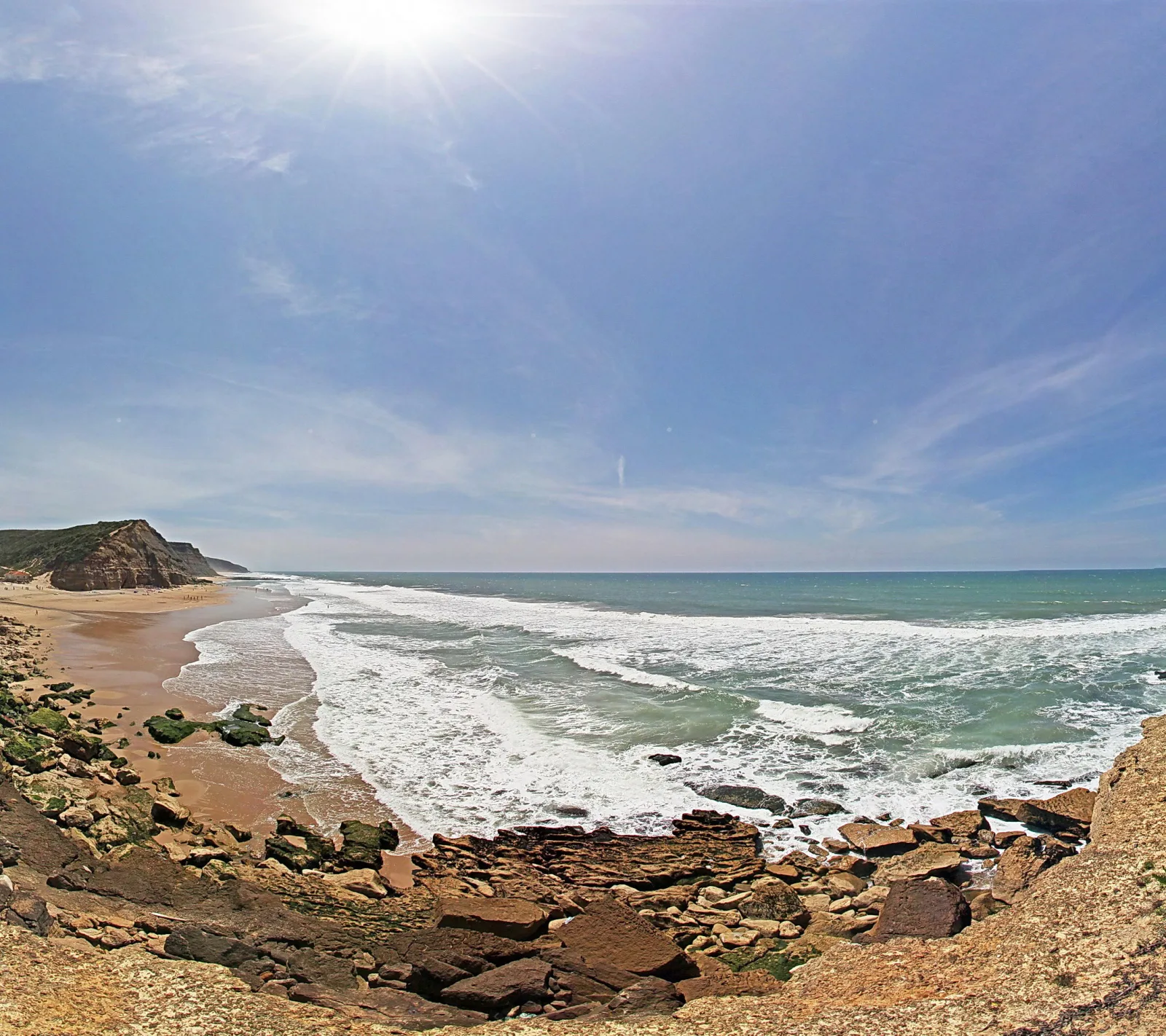 Felsige Küste mit Sandstrand, Brandung und Sonne über dem Meer unter blauem Himmel