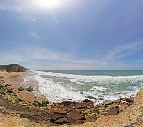 Felsige Küste mit Sandstrand, Brandung und Sonne über dem Meer unter blauem Himmel