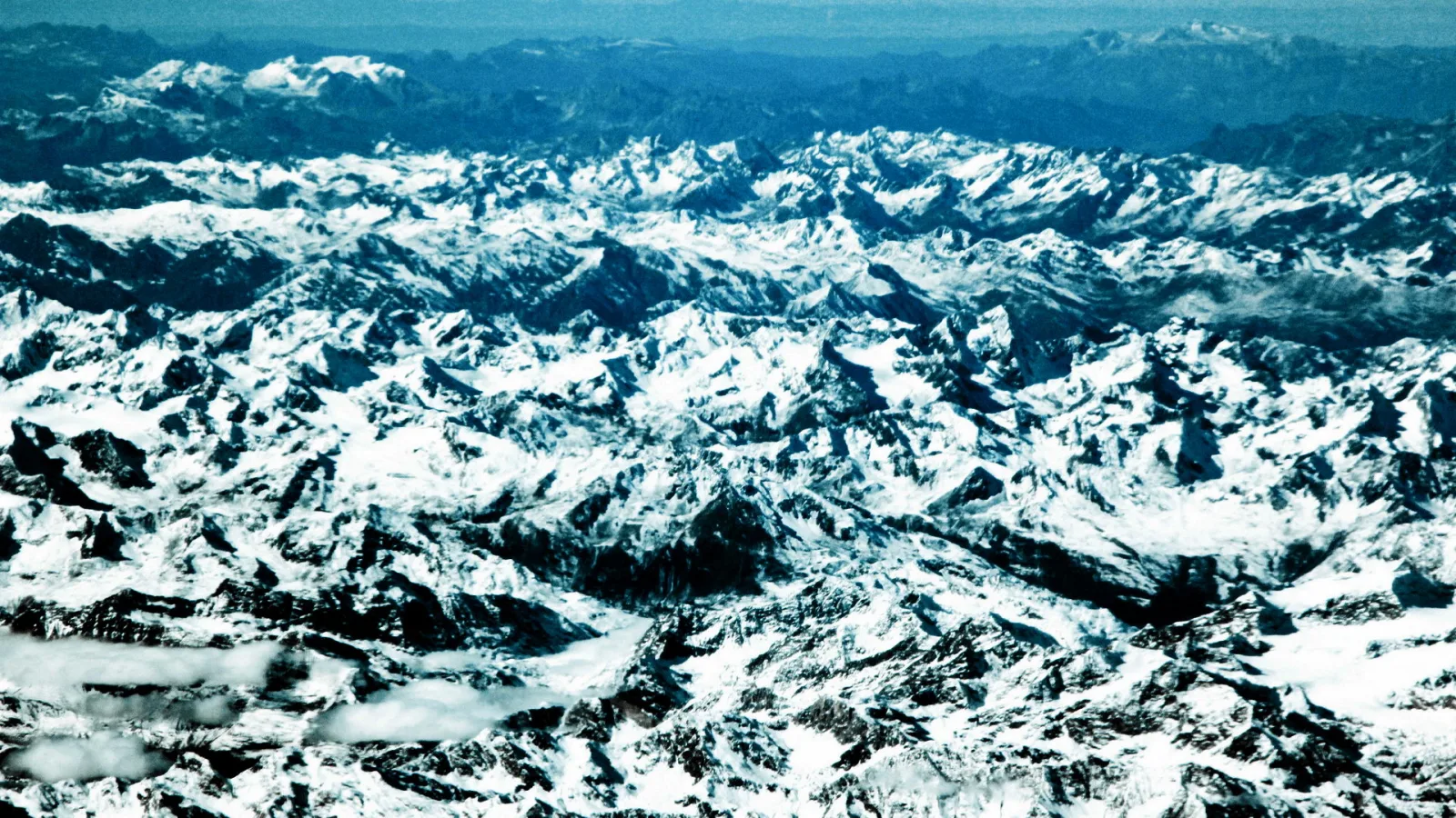 Luftaufnahme einer dicht zerklüfteten Gebirgskette mit schneebedeckten Gipfeln und dunklen Felsgraten, im Hintergrund blauer Dunst.