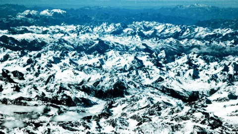 Luftaufnahme einer dicht zerklüfteten Gebirgskette mit schneebedeckten Gipfeln und dunklen Felsgraten, im Hintergrund blauer Dunst.