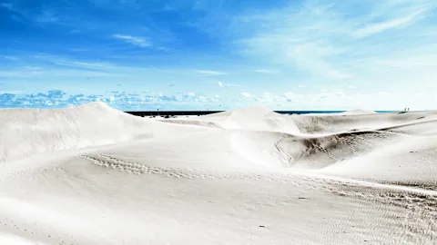 Helle, weiche Sanddünen unter blauem Himmel mit wenigen Wolken, dahinter ein dunkler Meereshorizont.