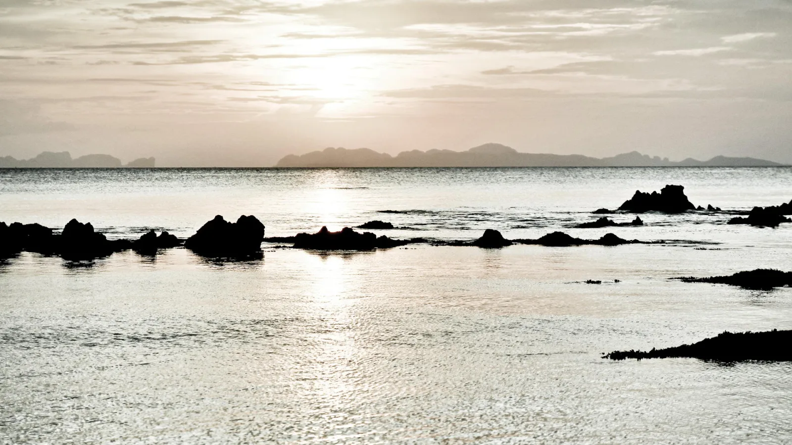Dunkle Felsen ragen aus ruhigem Meer, im Hintergrund zeichnen sich Inselketten am hellen, dämmrigen Horizont ab.