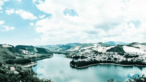 Panoramablick auf einen langgezogenen See, an dessen Ufer ein Dorf auf einem Hügel liegt, umgeben von grünen Wäldern und hellen Feldern unter weißen Wolken.