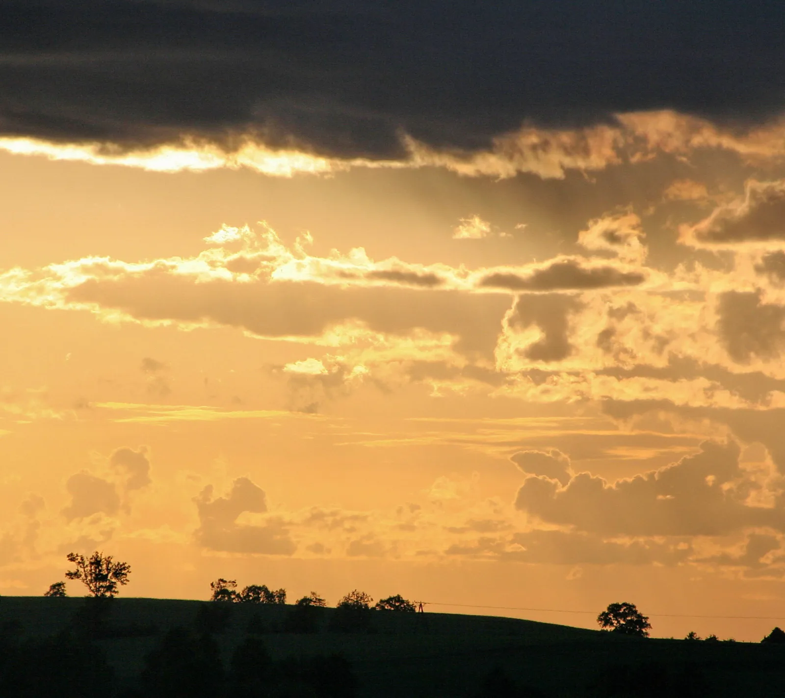 Sonnenuntergang über einer Hügellandschaft: dunkle Wolken oben, orangefarbener Himmel, dunkle Baumsilhouetten und eine Stromleitung am Horizont.