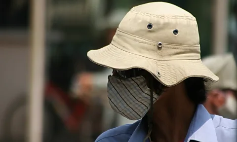 Person mit beigem Buschhut und gestreifter Stoffmaske vor unscharfem Hintergrund, im Profil