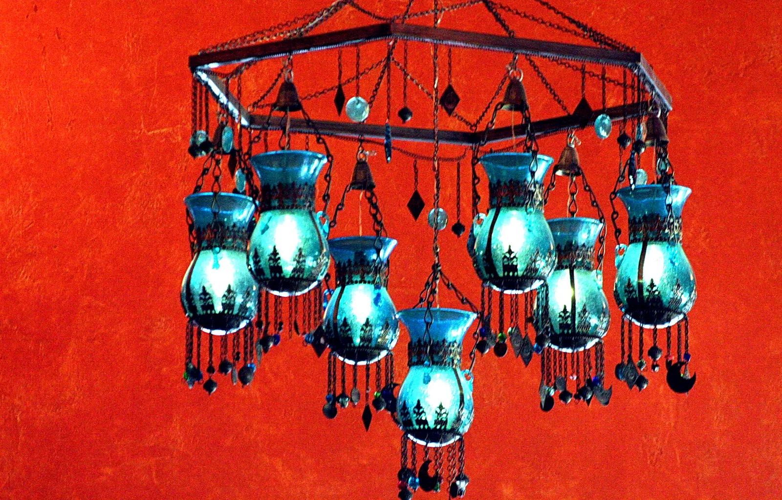 Hängende Kronleuchter aus türkisfarbenen Glasgefäßen mit dunklen Metallornamenten, Ketten und Glasperlen, vor einer leuchtend roten Wand