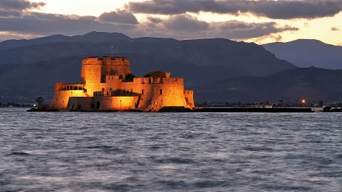 Beleuchtete Festung Bourtzi auf einer kleinen Insel im Wasser, dahinter Bergketten unter einem Abendhimmel mit Wolken.
