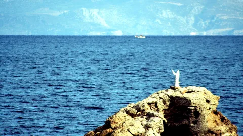 Weiße Jesusfigur mit erhobenen Armen auf einem Felsen über blauem Meer, im Hintergrund dunstige Küstenberge und ein kleines Boot.