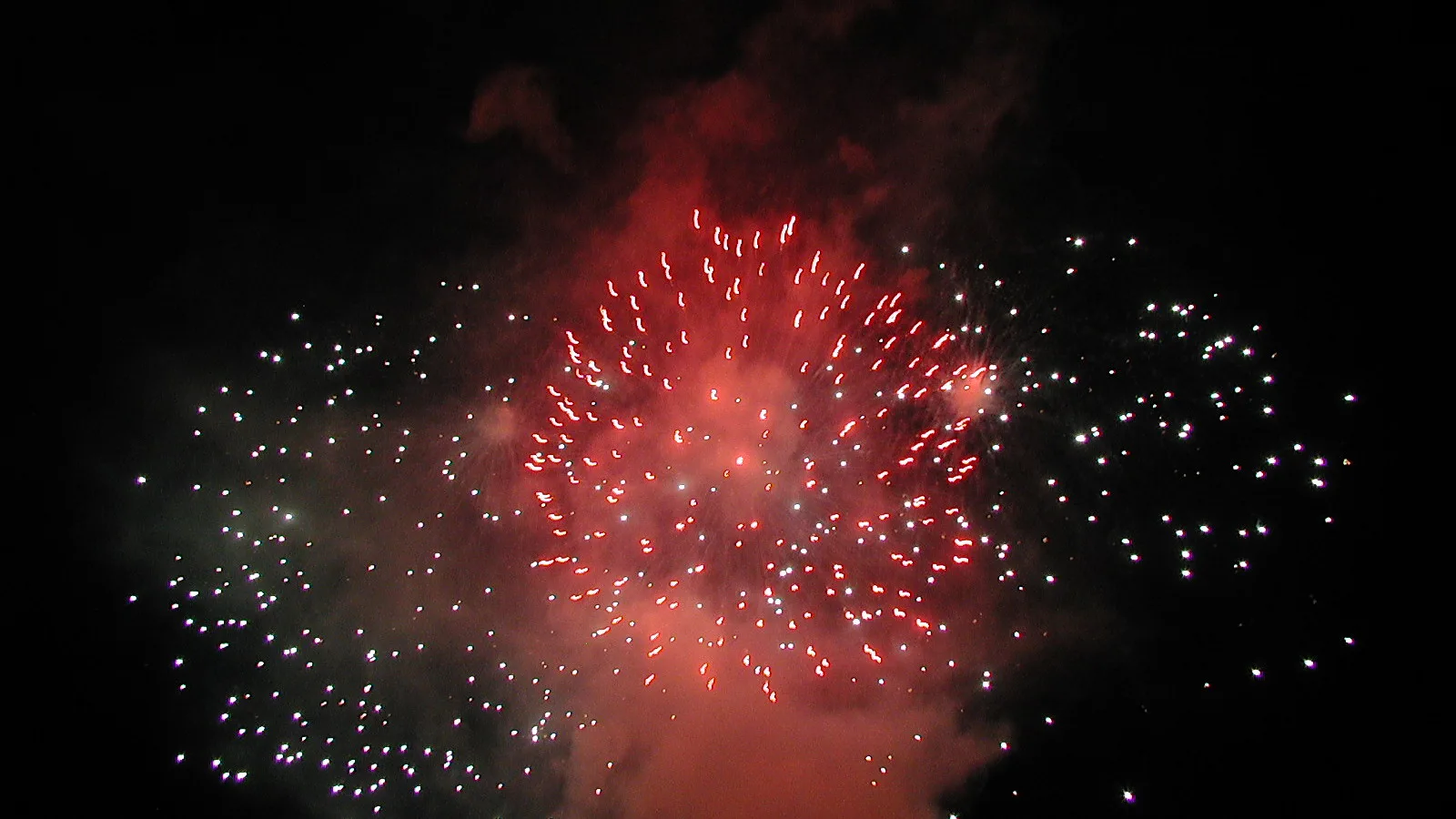 Ein rot leuchtendes Feuerwerk mit vielen weißen Funken vor dunklem Nachthimmel, umgeben von Rauchschwaden.