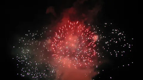 Ein rot leuchtendes Feuerwerk mit vielen weißen Funken vor dunklem Nachthimmel, umgeben von Rauchschwaden.