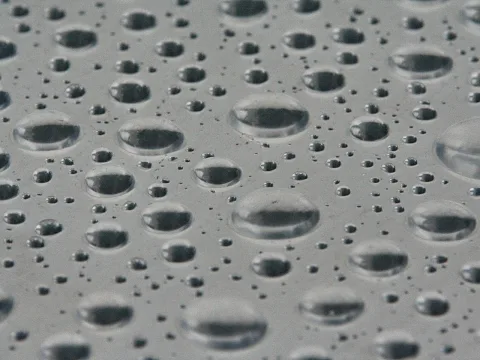 Viele Wassertropfen unterschiedlicher Größe liegen verteilt auf einer grauen, glatten Oberfläche und spiegeln die Umgebung.