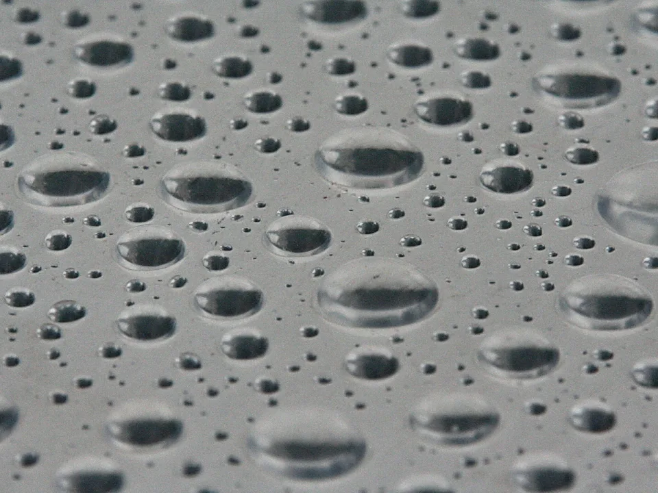 Viele Wassertropfen unterschiedlicher Größe liegen verteilt auf einer grauen, glatten Oberfläche und spiegeln die Umgebung.
