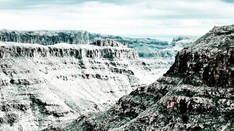 Schroffe, geschichtete Felsplateaus und eine tiefe Schlucht unter einem bewölkten Himmel, in kühlen, entsättigten Grau- und Blaugrüntönen.