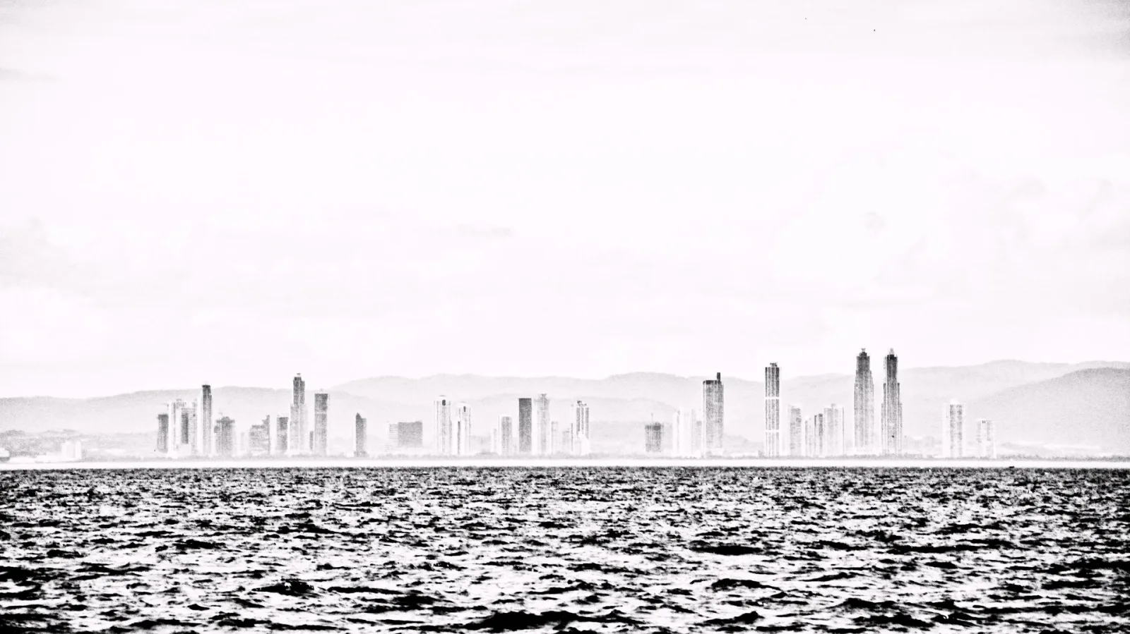 Schwarzweiß-Foto einer Hochhaus-Skyline am Horizont über einer bewegten Wasserfläche, darüber heller, diesiger Himmel.