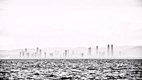 Schwarzweiß-Foto einer Hochhaus-Skyline am Horizont über einer bewegten Wasserfläche, darüber heller, diesiger Himmel.