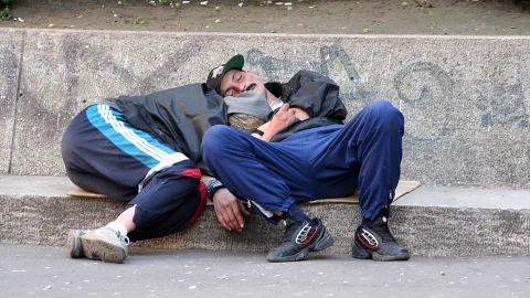 Ein Mann mit Basecap und Jogginghose schläft auf einem Betonrandstein, im Hintergrund eine graue Mauer mit Graffiti.