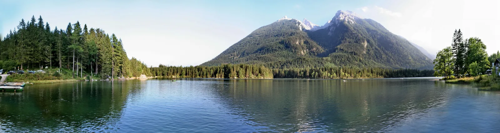 Panoramablick auf einen ruhigen Bergsee mit dunkelgrünem Nadelwald am linken Ufer und einem steilen, bewaldeten Berg mit felsigen Gipfeln im Hintergrund. Im Vordergrund ein kleiner Steg und Boote am Ufer.