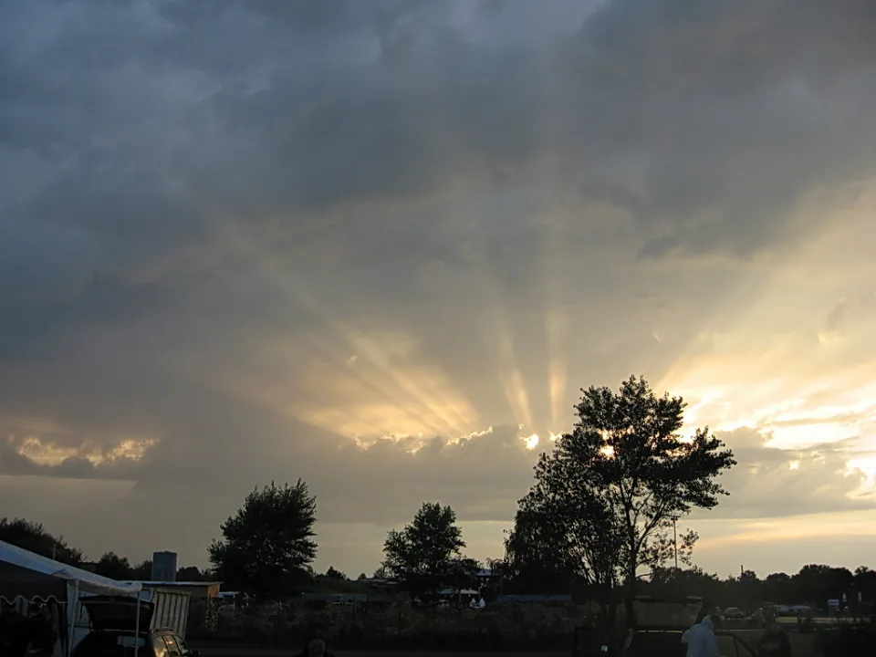 Sonnenstrahlen brechen durch eine dunkle Wolkendecke über silhouettierten Bäumen und einem Zeltdach am Abend.