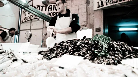 Ein Fischverkäufer mit Brille und weißer Schürze steht hinter einem großen Haufen Miesmuscheln, davor liegen Fische auf Eis. Im Hintergrund hängen Schilder mit der Aufschrift La Pescheria, links arbeitet ein zweiter Mann.