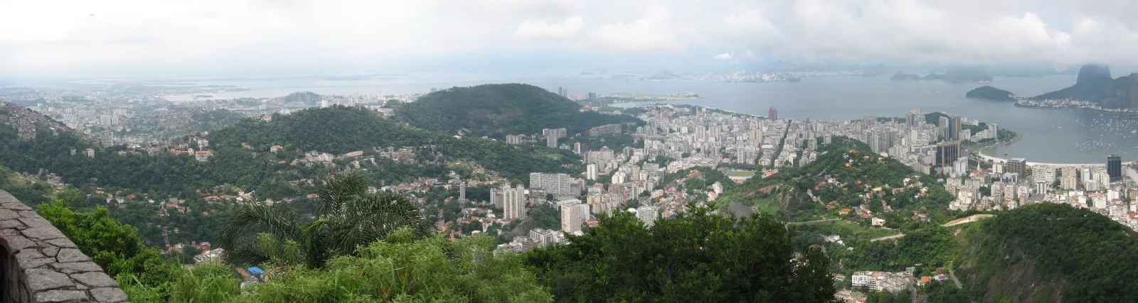 Panoramablick über die dicht bebaute Stadt Rio de Janeiro mit grünen Hügeln im Vordergrund, einer breiten Bucht und Hochhäusern am rechten Rand unter bewölktem Himmel.