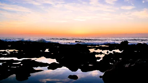 Sonnenuntergang über dem Meer, weiße Wellen brechen am Ufer, im Vordergrund dunkle Felsen mit flachen Wasserbecken, die den orangefarbenen Himmel spiegeln.