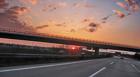 Eine mehrspurige Autobahnbrücke mit Geländer verläuft quer über das Bild. Darunter fahren Autos auf der Autobahn, rechts ist ein Verkehrsschild über der Fahrbahn zu sehen. Der Himmel ist orange und rosa mit Wolken, die Sonne steht tief am Horizont.