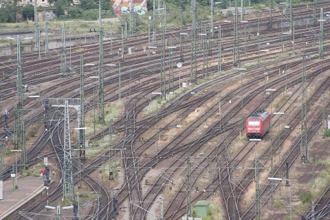 Weitläufiges Gleisfeld mit Oberleitungsmasten, rechts fährt eine rote Lokomotive durch das Gewirr aus Schienen.