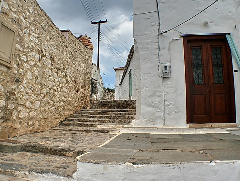 Steile Steintreppe zwischen einer Bruchsteinmauer links und einem weiß getünchten Haus mit brauner Doppeltür rechts, darüber bewölkter Himmel und eine Stromleitung.
