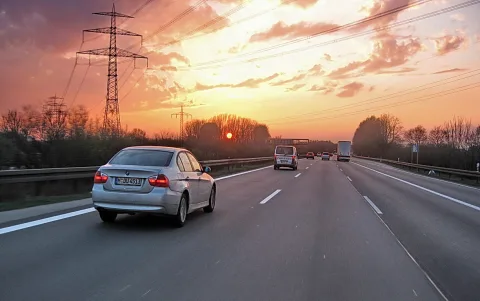 Ein silberner BMW fährt bei Sonnenuntergang über eine Autobahn, links und rechts stehen Bäume, im Hintergrund ragen Strommasten in den rosa Himmel.
