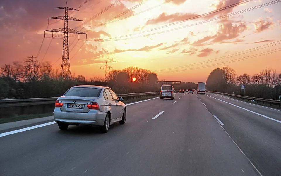 Ein silberner BMW fährt bei Sonnenuntergang über eine Autobahn, links und rechts stehen Bäume, im Hintergrund ragen Strommasten in den rosa Himmel.