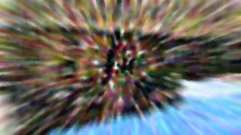 Farbig radial verwischte Grün- und Rosatöne strahlen von einem dunklen Mittelpunkt aus, unten rechts ist blauer Himmel zu sehen.