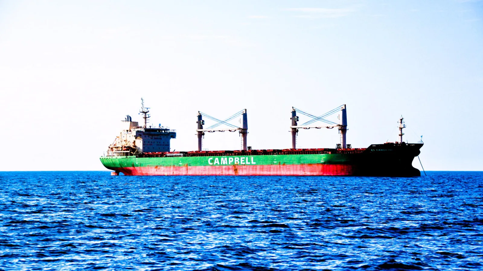Ein grün und rot gestrichenes Frachtschiff mit der Aufschrift CAMPBELL und Kränen fährt über ein blaues, leicht bewegtes Meer unter hellem Himmel.