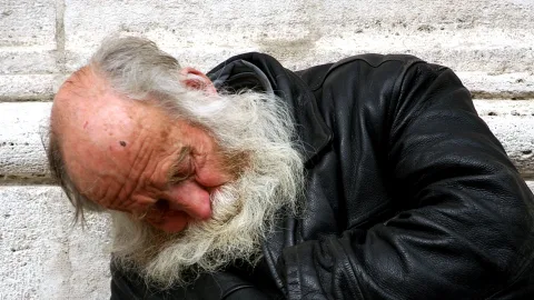 Ein alter Mann mit grauem Bart und kahlem Kopf lehnt in einer schwarzen Lederjacke mit gesenktem Kopf vor einer hellen Steinmauer.