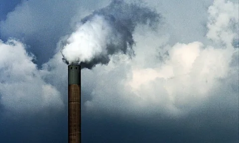 Ein schmaler Schornstein ragt von unten ins Bild, aus seiner Spitze steigt weißer Rauch in einen grau bewölkten Himmel.