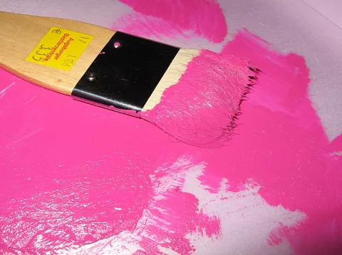 Ein Flachpinsel mit Holzgriff und pinker Farbe in den Borsten liegt auf einer Fläche mit breiten pinken Farbstrichen.