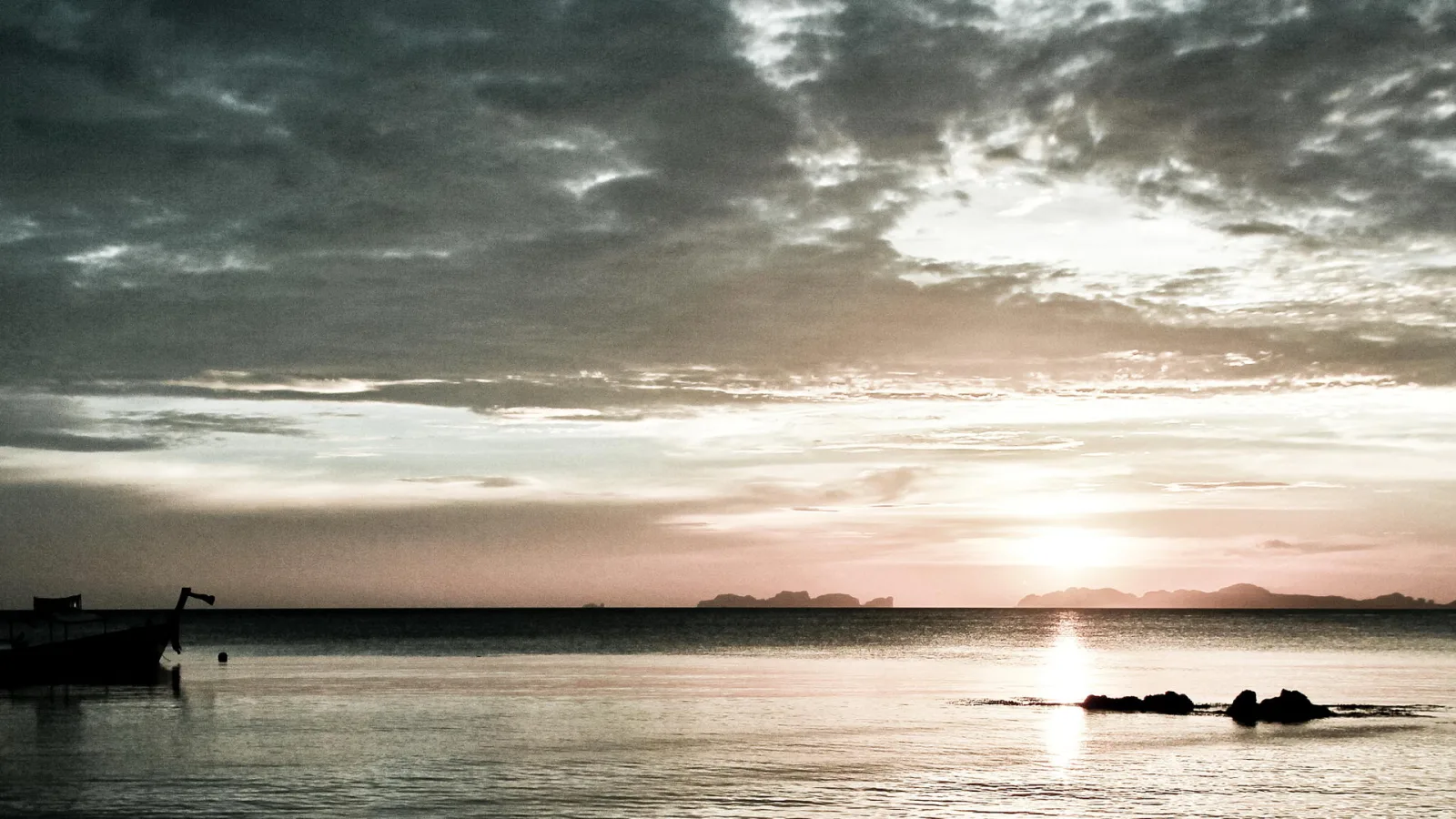 Ein kleines Boot liegt links am Rand einer ruhigen Wasserfläche, rechts ragen Felsen aus dem Meer, am Horizont zeichnen sich Inseln ab, und die Sonne sinkt hinter dunklen Wolken.