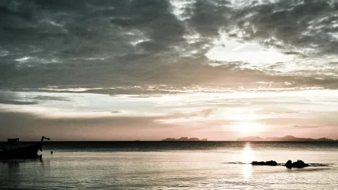 Ein kleines Boot liegt links am Rand einer ruhigen Wasserfläche, rechts ragen Felsen aus dem Meer, am Horizont zeichnen sich Inseln ab, und die Sonne sinkt hinter dunklen Wolken.